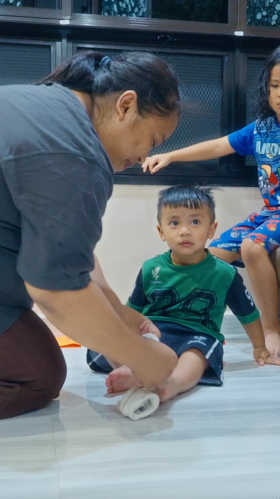 A helper putting on a toddler's socks at home, with the other children close by