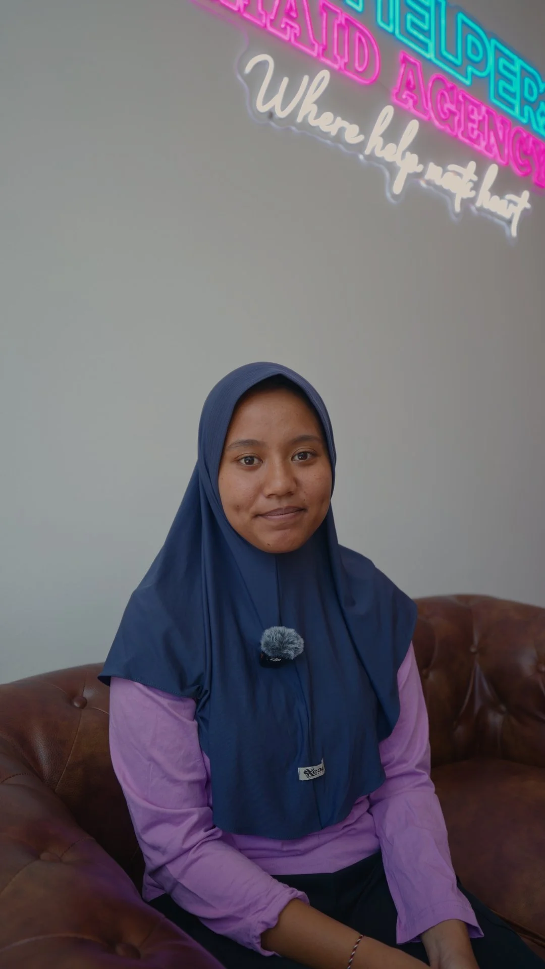 A woman in a hijab seated on the sofa beneath the neon sign at the Jaz Helpers office